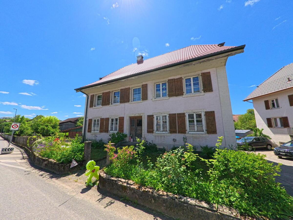 Ein älteres Wohnhaus mit braunen Fensterläden und einem blühenden Vorgarten steht an einer verkehrsberuhigten Strasse in Basse-Vendline (2944, Kanton Jura).
