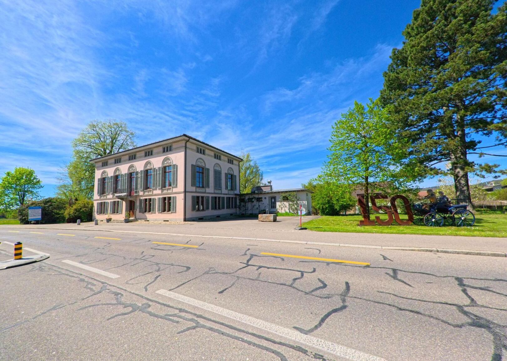 In Bassersdorf (8303, Kanton Zürich) steht ein historisches Gebäude mit grossen Rundbogenfenstern an einer Hauptstrasse, daneben erinnert eine grosse 