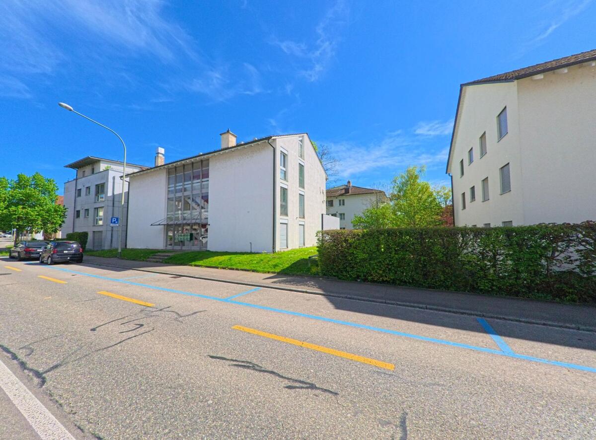 Entlang einer Durchgangsstrasse in Bassersdorf (8303, Kanton Zürich) stehen mehrere moderne Wohnhäuser mit grossen Glasflächen und Parkplätzen am Strassenrand.