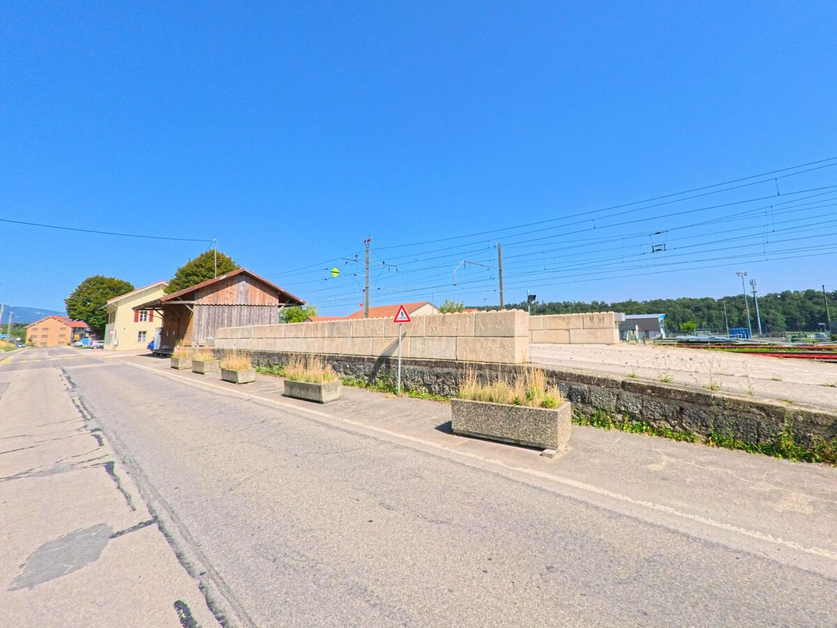 Am Bahnhofsgelaende von Baulmes (1446, Kanton Waadt) verlaeuft eine Strasse entlang der Gleise, begrenzt von Mauern und alten Nebengebaeuden.