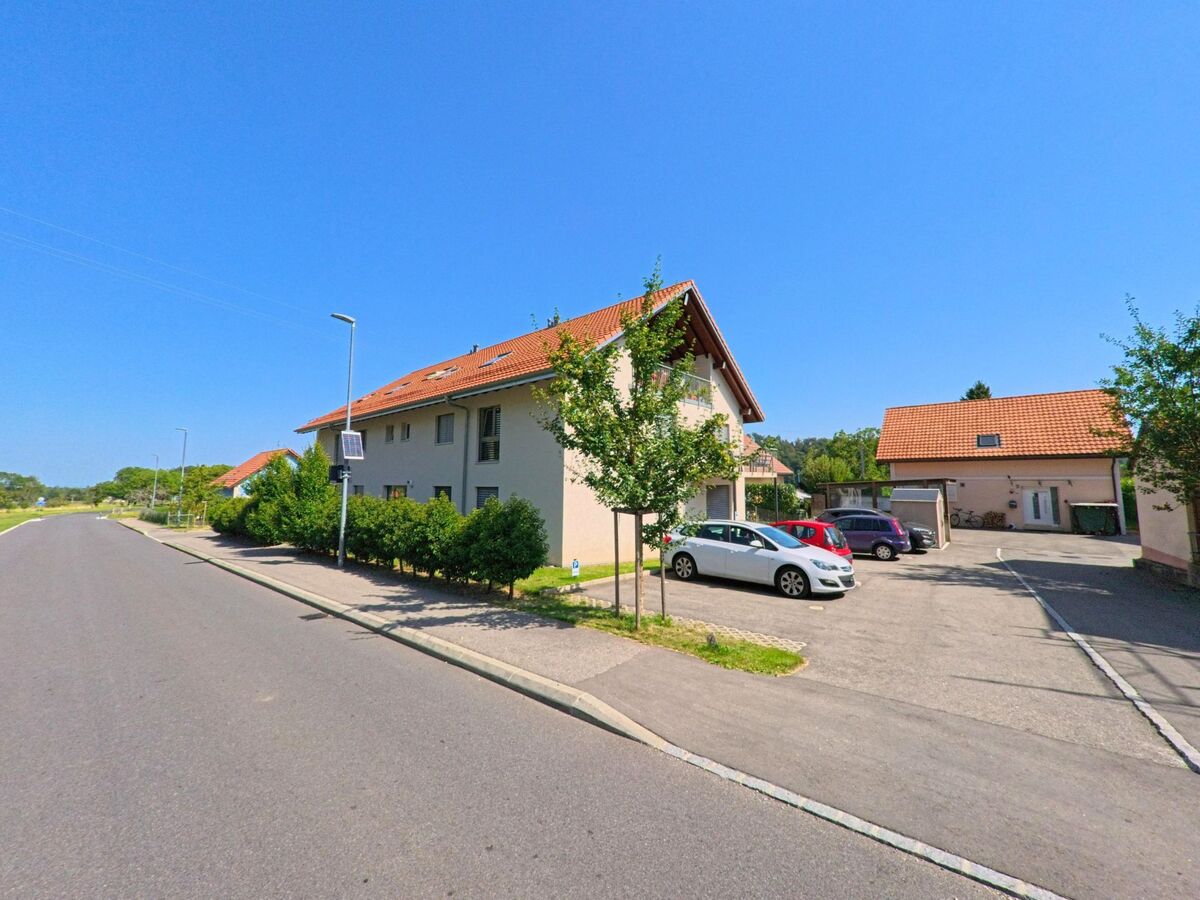 Ein modernes Wohnhaus mit rotem Dach und angrenzendem Parkplatz steht in Baulmes (1446, Kanton Waadt) an einer breiten Strasse mit jungen Baeumen.