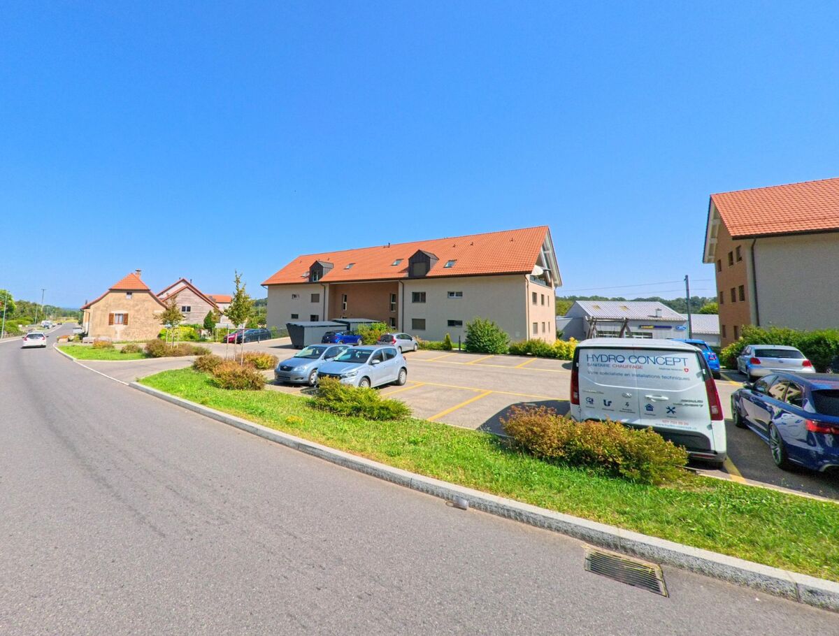 An einer breiten Strasse in Baulmes (1446, Kanton Waadt) stehen moderne Wohngebaeude mit roten Daechern, davor befinden sich Parkplaetze mit mehreren Fahrzeugen.