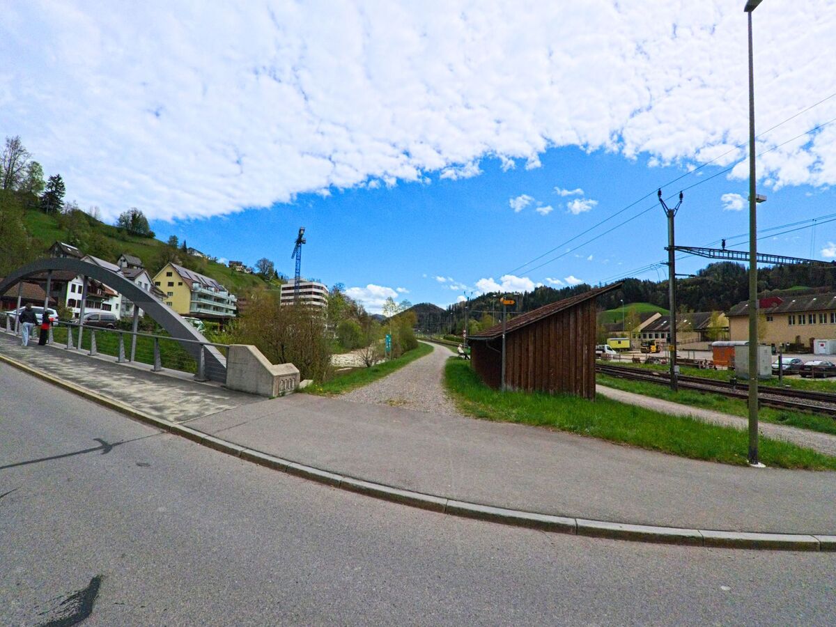 In Bauma im Kanton Zürich, PLZ 8494, führt ein Fussweg entlang der Bahngleise mit Blick auf die markante Brücke über die Töss und umliegende Wohnhäuser.