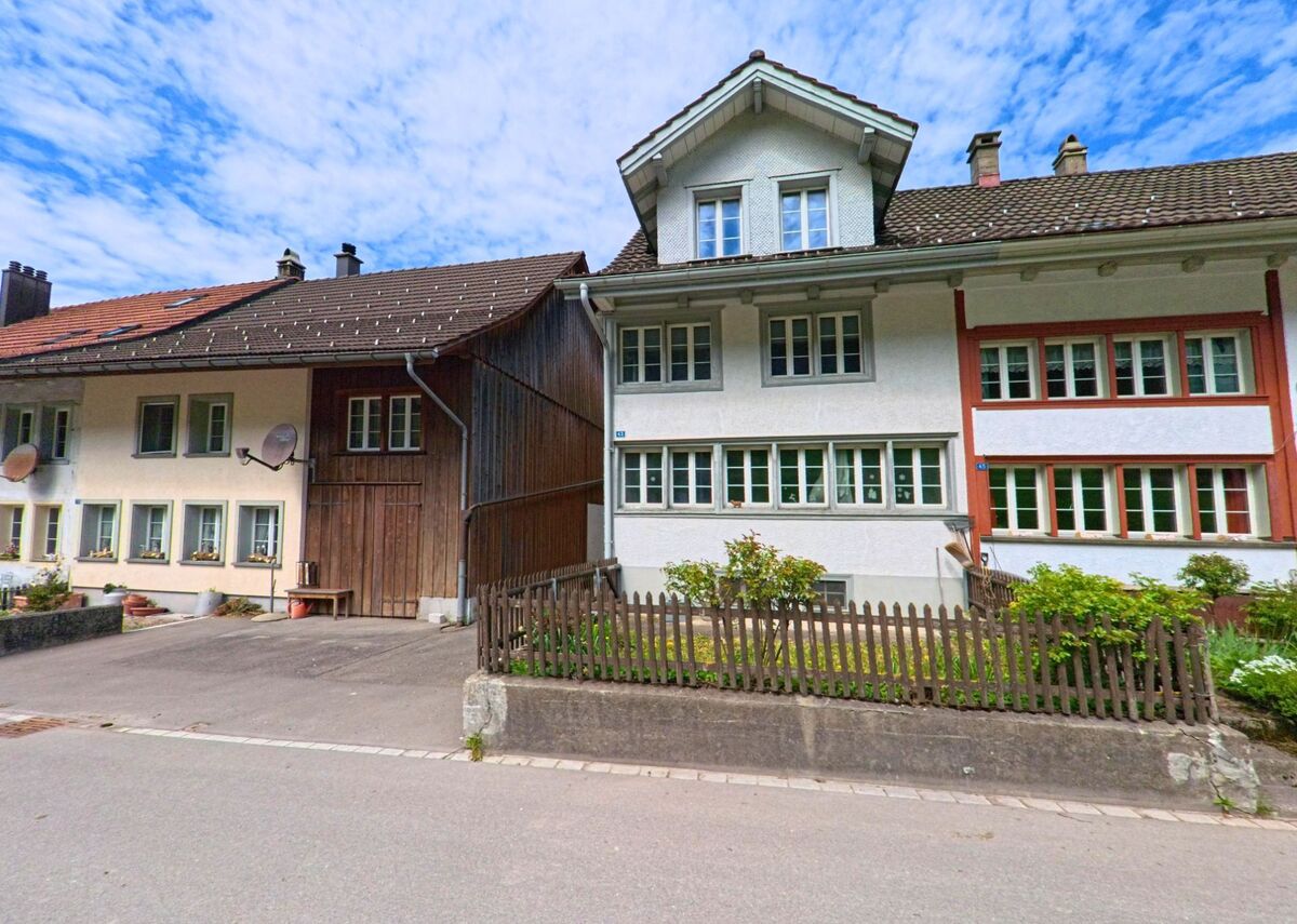  In Bauma (8494, Kanton Zürich) stehen traditionell gebaute Wohnhäuser mit gepflegten Fenstern und Vorgärten entlang einer ruhigen Quartierstrasse unter leicht bewölktem Himmel.