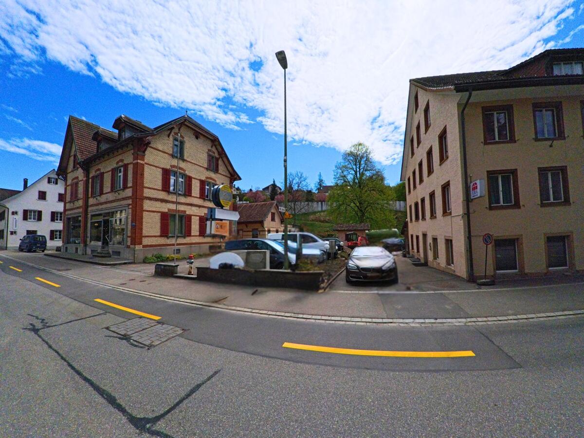 Zentral in Bauma im Kanton Zürich, PLZ 8494, stehen ein Geschäftshaus mit rotem Fensterladen und ein Wohnhaus gegenüber eines kleinen Parkplatzes an einer Hauptstrasse.