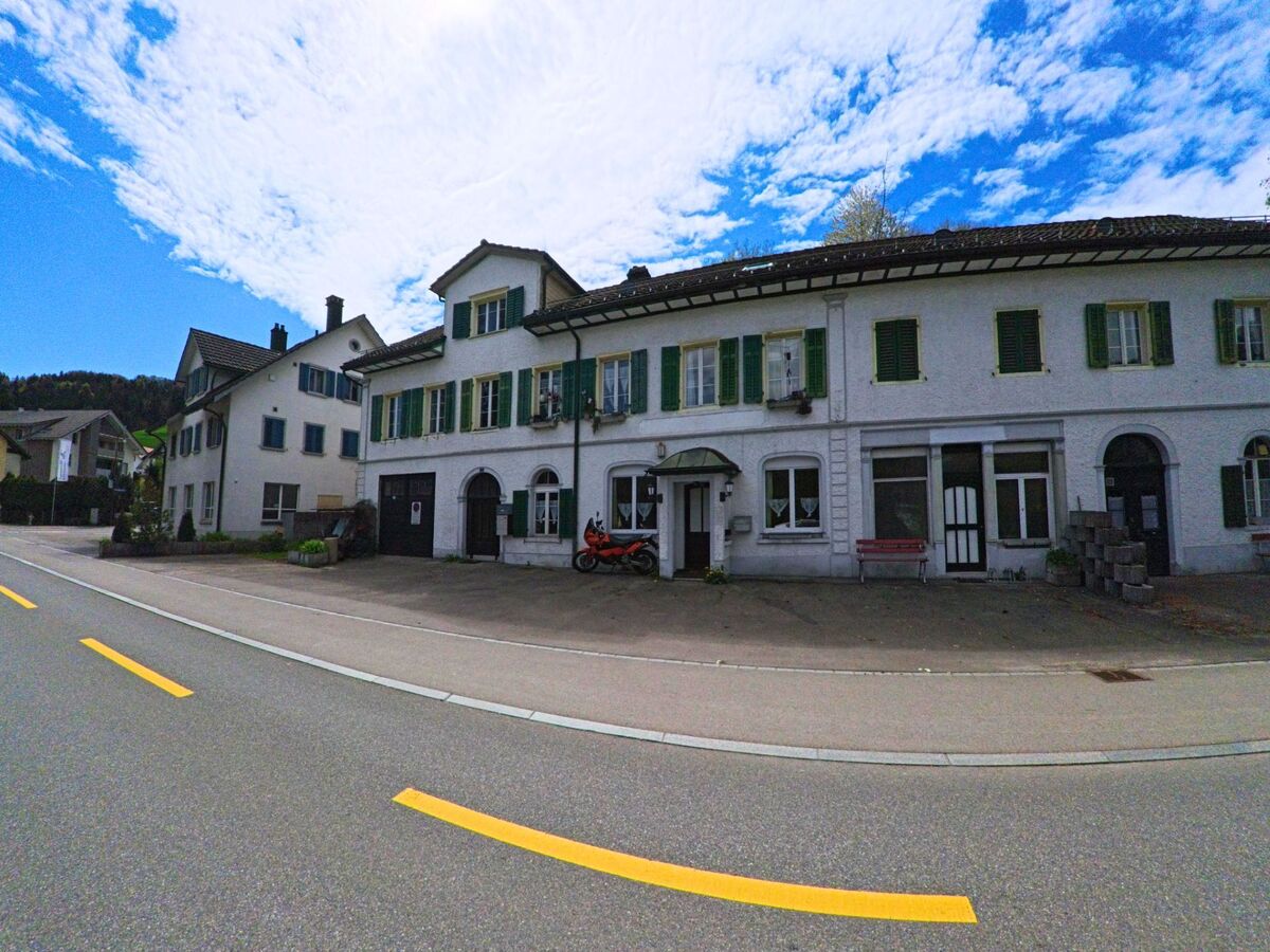 Entlang der Hauptstrasse in Bauma im Kanton Zürich, PLZ 8494, stehen mehrere Häuser mit grünen Fensterläden und einer roten Sitzbank vor einem ehemaligen Geschäft.