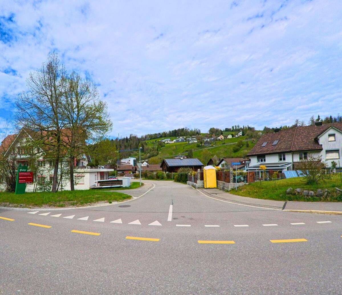 Zentral in Bauma im Kanton Zürich, PLZ 8494, stehen ein Geschäftshaus mit rotem Fensterladen und ein Wohnhaus gegenüber eines kleinen Parkplatzes an einer Hauptstrasse.