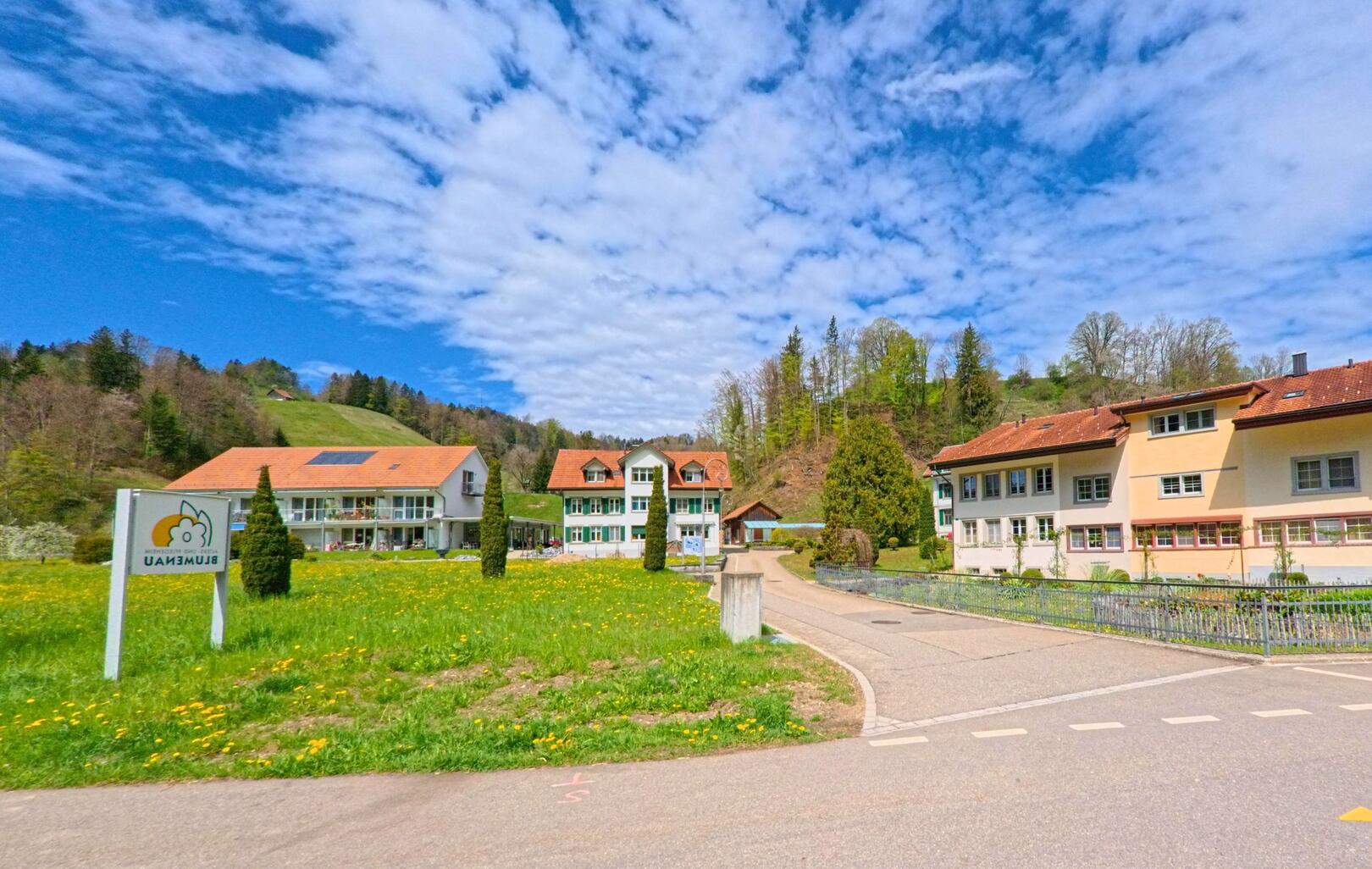 In Bauma (8494, Kanton Zürich) liegt das Gelände der Stiftung Blumenau mit mehreren Gebäuden inmitten einer Frühlingswiese am Waldrand unter blauem Himmel.