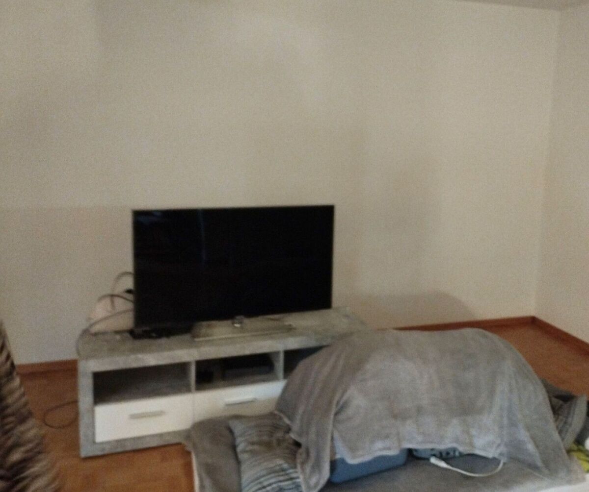 In einem Wohnzimmer in Bauma (8494, Kanton Zürich) steht ein grosser Fernseher auf einem TV-Möbel vor einer weissen Wand, daneben ein Sofa mit einer Decke.