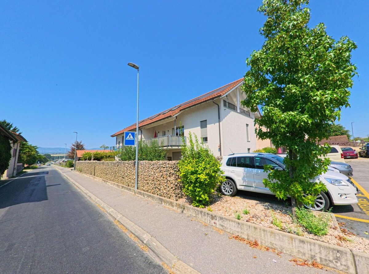 Ein Wohnhaus in Bavois (1372, Kanton Waadt) mit heller Fassade und Balkon liegt an einer Strasse neben einem Parkplatz.