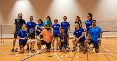 In Baar (6314, Kanton Zug) posiert eine grosse Badmintongruppe in einer Sporthalle vor dem Netz, waehrend die Spielerinnen und Spieler lachend mit ihren Schlaegern knien oder stehen.