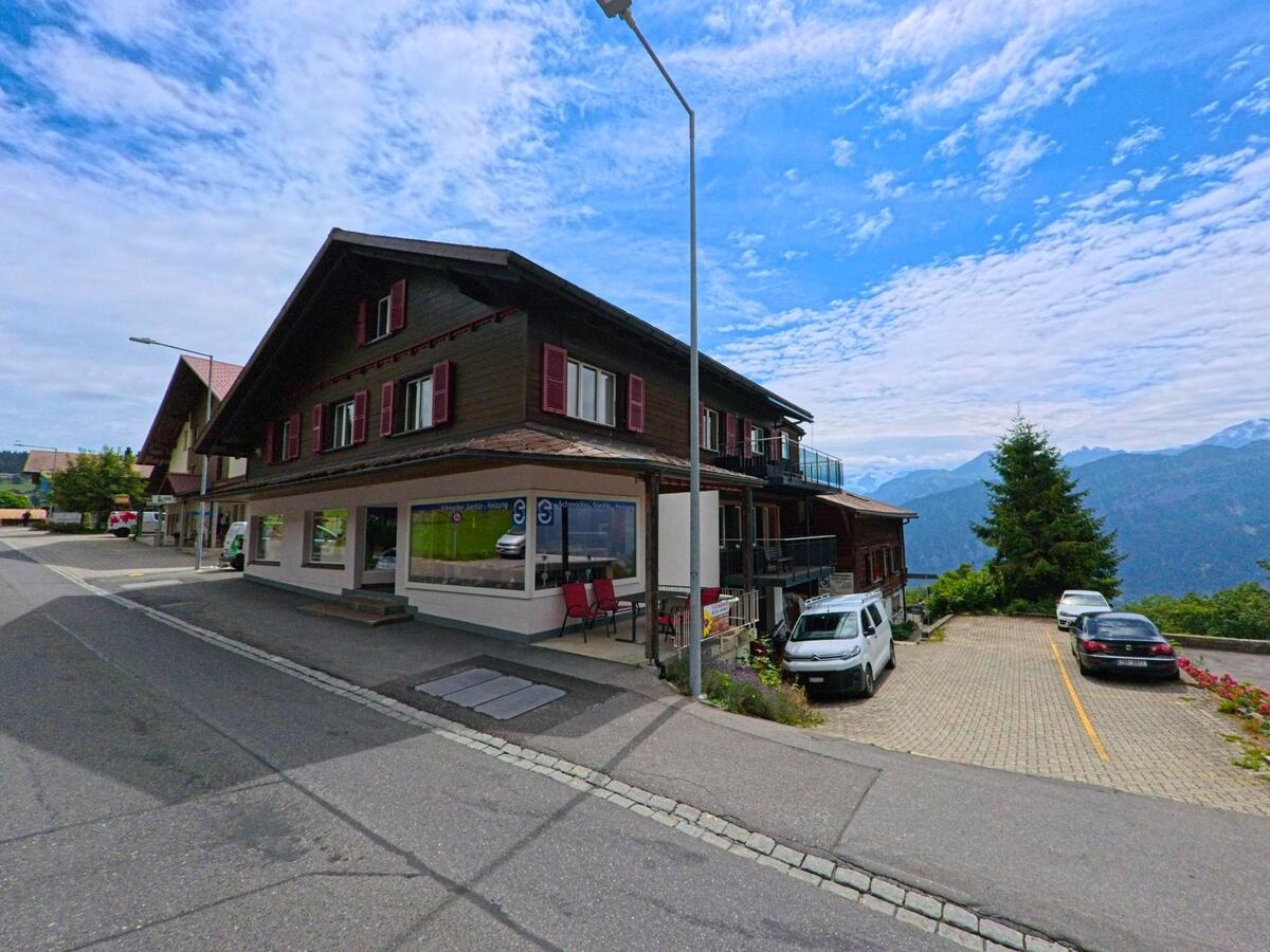 Ein Gebäude mit Ladengeschäft und roten Fensterläden steht in Beatenberg (3803, Kanton Bern) an einer Anhöhe mit Parkplatz und Blick auf die umliegenden Berge.