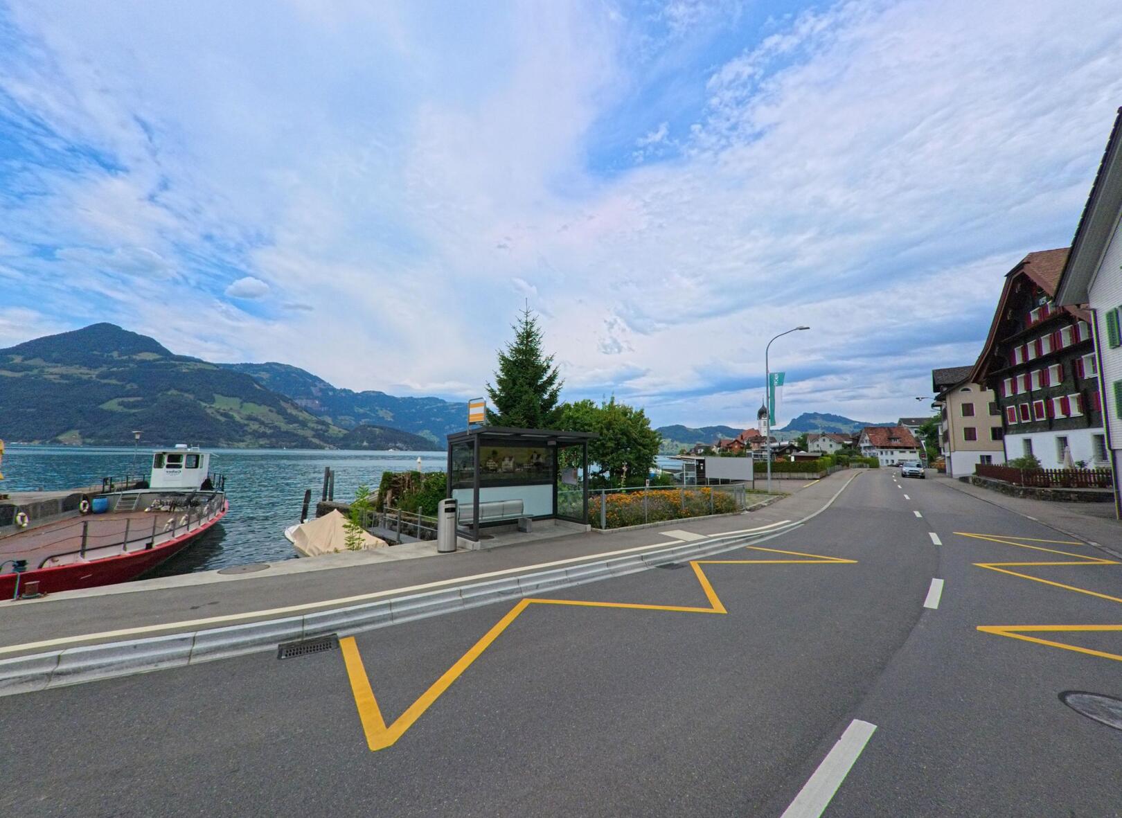 In Beckenried (6375, Kanton Nidwalden) liegt eine Bushaltestelle direkt am Vierwaldstättersee neben einem Anlegesteg mit einem Schiff und Blick auf die umliegenden Berge.