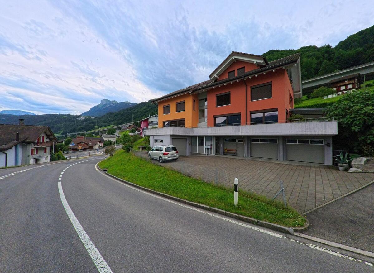 In Beckenried (6375, Kanton Nidwalden) steht ein oranger Wohnbau mit Garagen an einer kurvigen Hangstrasse mit Blick auf ein markantes Bergmassiv im Hintergrund.