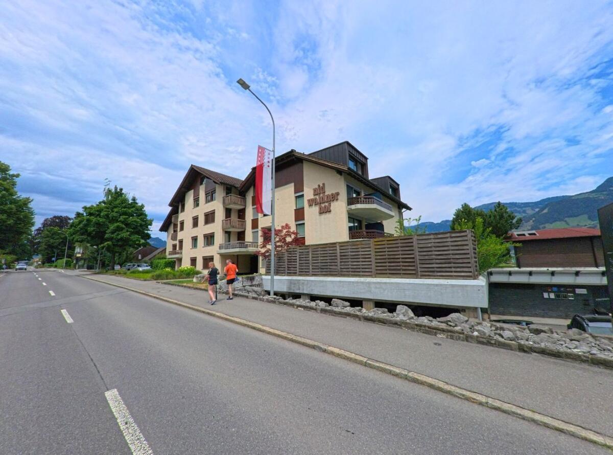 In Beckenried (6375, Kanton Nidwalden) gehen zwei Personen auf einem Gehweg entlang eines modernen Wohnhauses mit dem Schriftzug „nid wäldner hof“ an einer breiten Strasse.