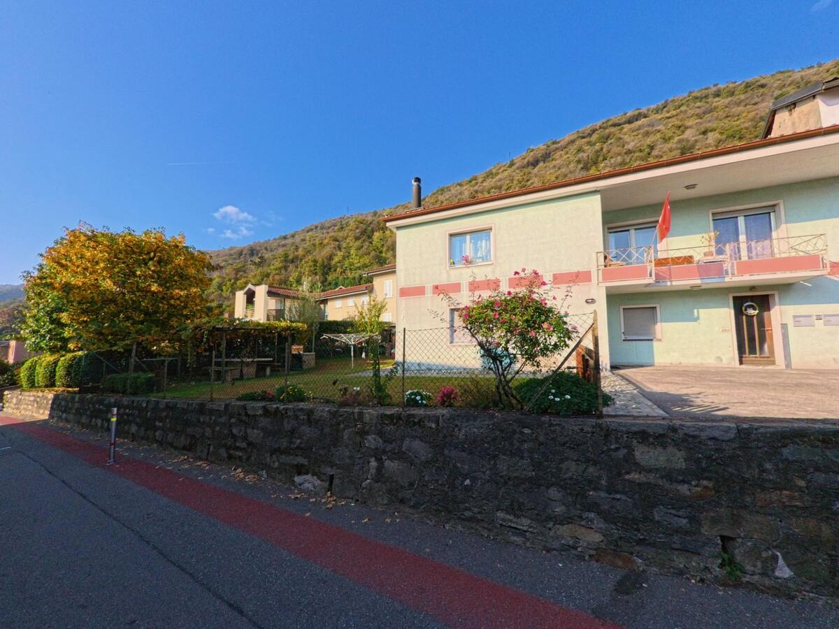 Entlang einer Wohnstrasse in Bedano (6930, Kanton Tessin) stehen Mehrfamilienhaeuser mit Garten hinter einer niedrigen Steinmauer am bewaldeten Hang.