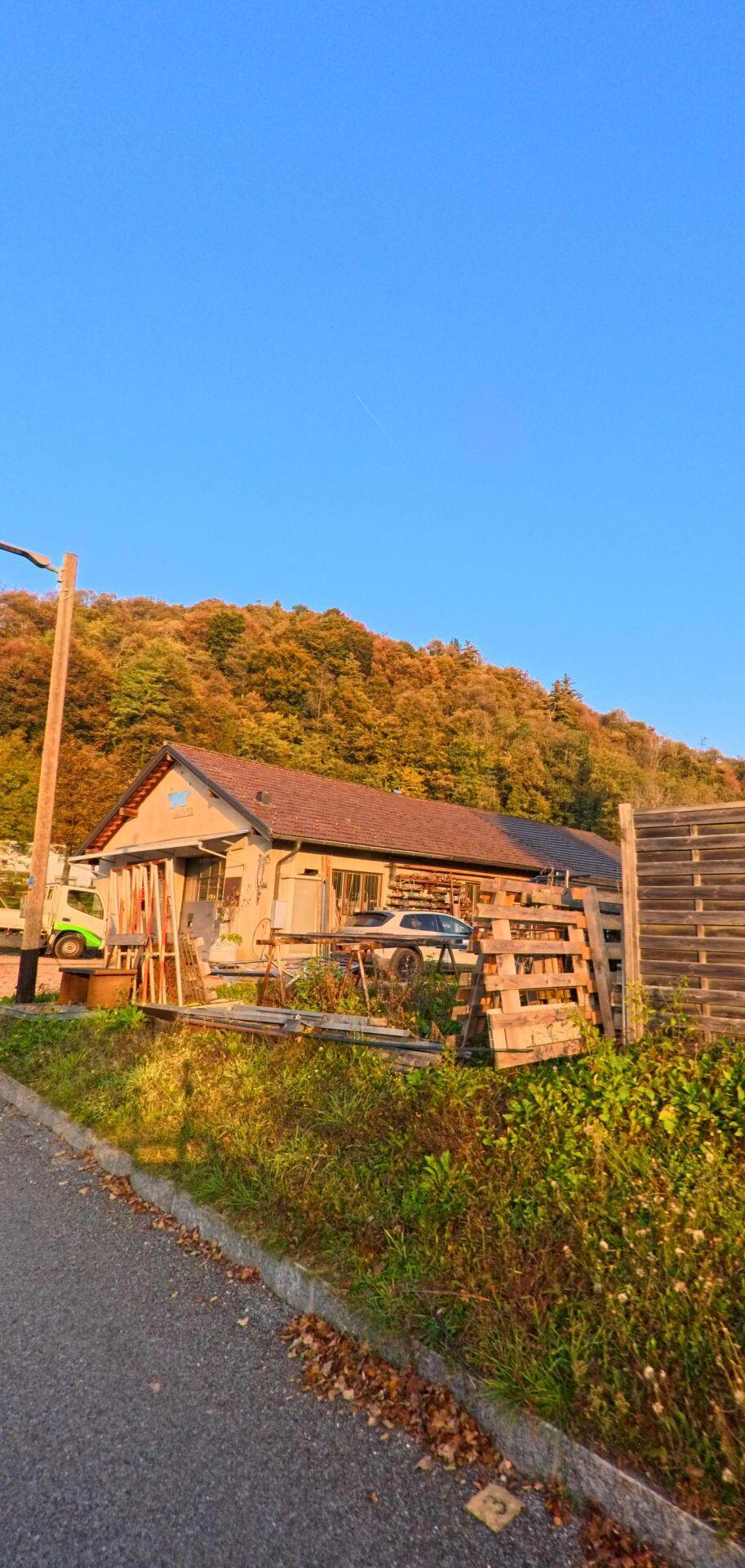 Am Rand einer schmalen Strasse ist in Bedigliora (6981, Kanton Tessin) ein einfaches Gewerbegebaeude mit Holzlager und Fahrzeugen vor einem herbstlich gefaerbten Waldhang zu sehen.