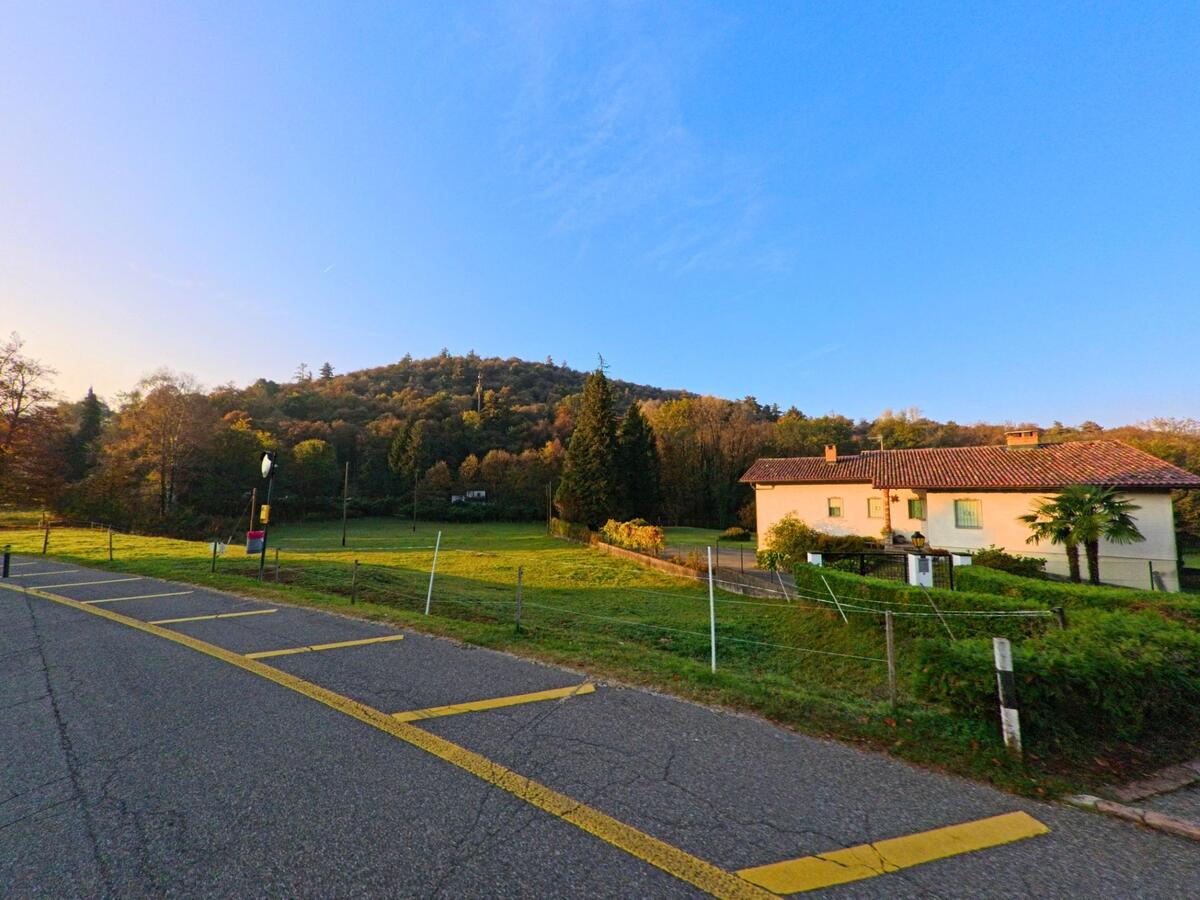 Neben markierten Parkfeldern erstreckt sich in Bedigliora (6981, Kanton Tessin) eine offene Gruenflaeche mit Wohnhaus und Blick auf bewaldete Huegel.