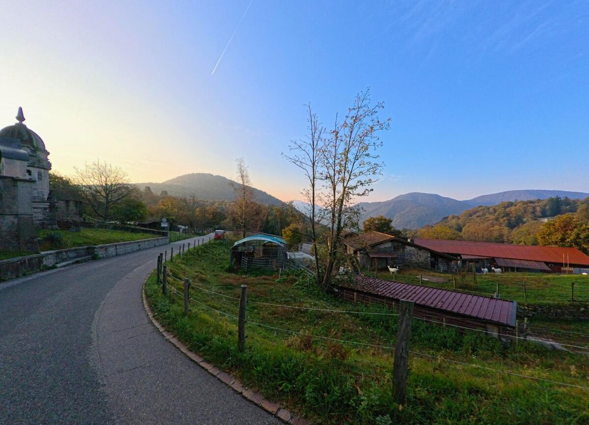 Eine sanft geschwungene Strasse fuehrt in Bedigliora (6981, Kanton Tessin) an einer kleinen Kapelle, Weideflaechen und landwirtschaftlichen Gebaeuden mit Blick auf die umliegenden Huegel vorbei.