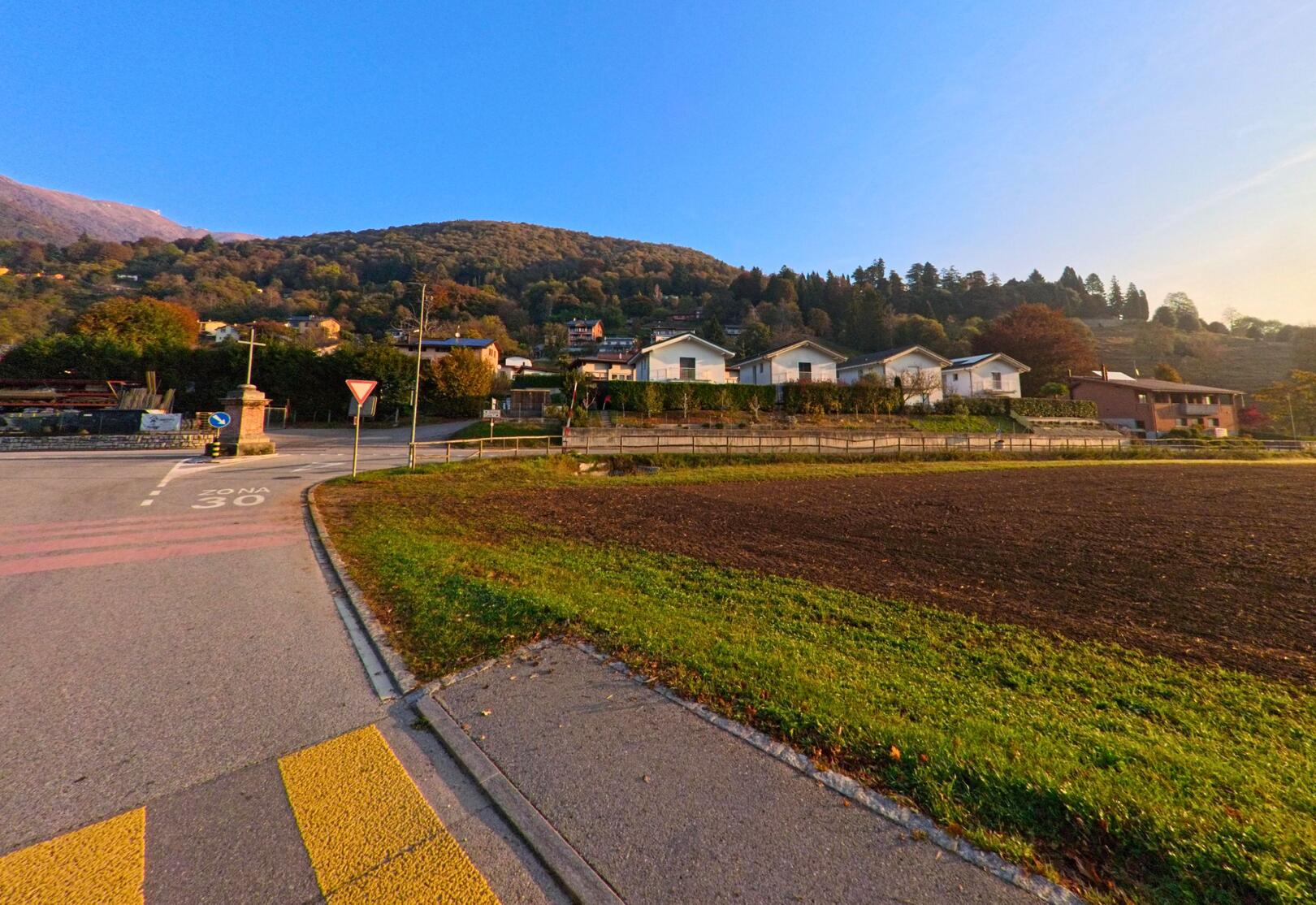 Am Rand einer Tempo-30-Zone zeigt sich in Bedigliora (6981, Kanton Tessin) eine Strassenkreuzung mit Wohnhaeusern, Ackerflaeche und bewaldetem Hang im Hintergrund.