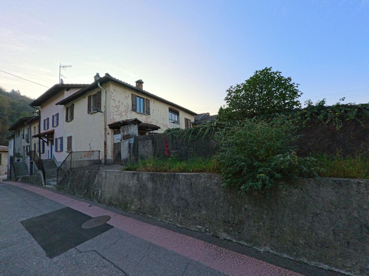 Entlang einer ruhigen Wohnstrasse stehen in Bedigliora (6981, Kanton Tessin) mehrere aeltere Hauser mit Fassaden in unterschiedlichem Zustand hinter einer niedrigen Mauer.