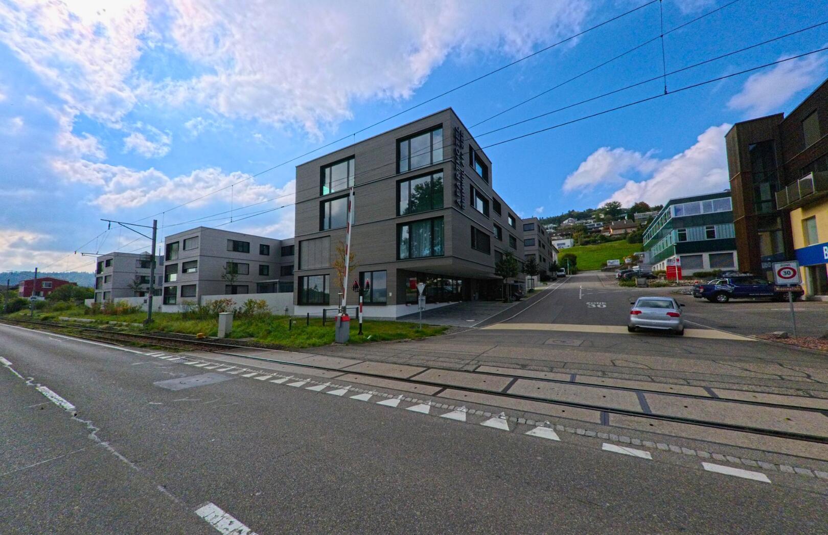 In Beinwil am See (5712, Kanton Aargau) führt eine Strasse mit Bahnübergang an modernen Wohngebäuden vorbei, die sich an einem sanft ansteigenden Hang befinden.
