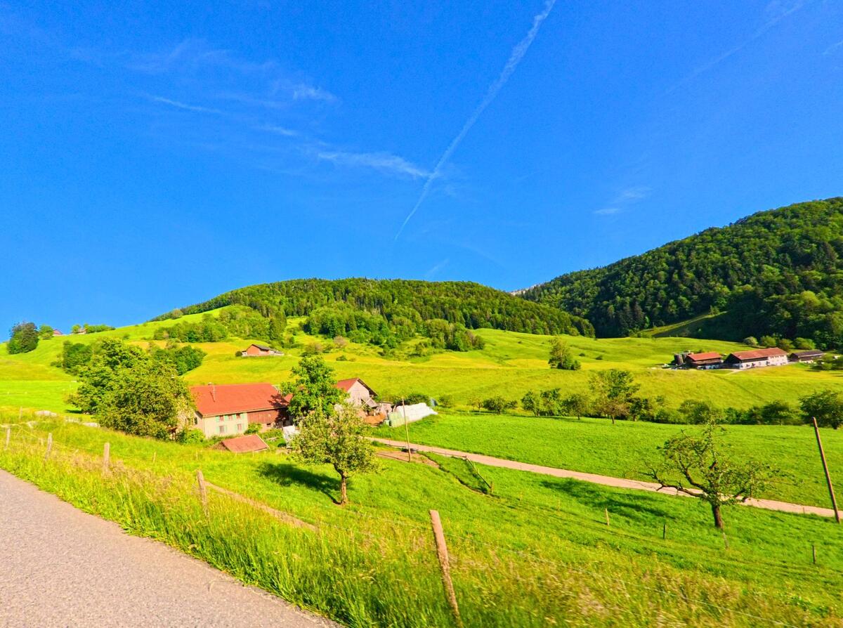 In Beinwil_(SO) im Kanton Solothurn, PLZ 4229, breitet sich eine hügelige Kulturlandschaft mit verstreuten Bauernhöfen und Wiesen unter klarem Himmel aus.