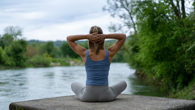 Eine Frau sitzt entspannt in Meditationshaltung am Flussufer in Schönenwerd (5012, Kanton Solothurn) und streckt die Arme hinter dem Kopf in ruhiger Umgebung.
