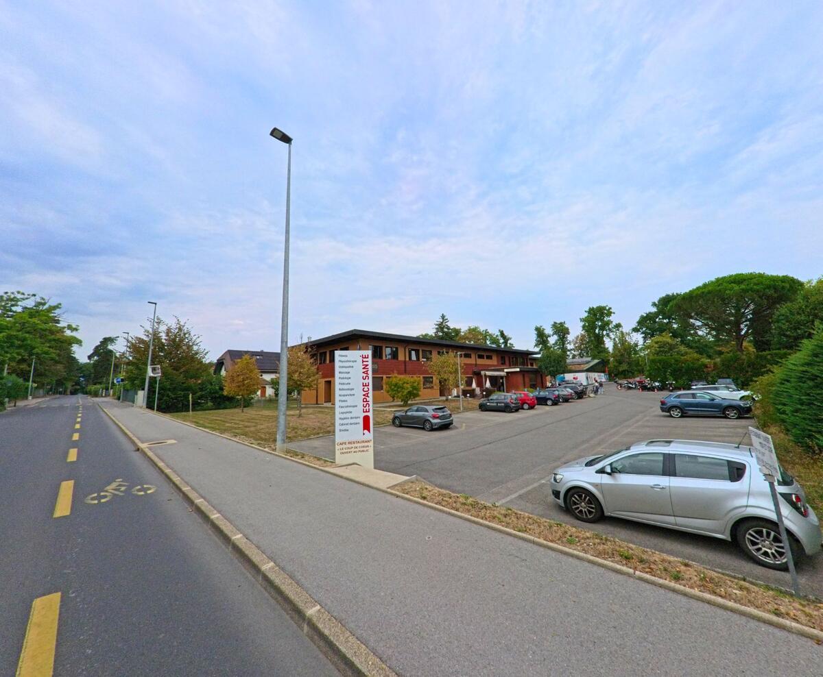 Vor einem Gesundheitszentrum mit grossem Parkplatz in Bellevue (1293, Kanton Genf) stehen zahlreiche Autos, daneben fuehrt eine Strasse mit Radstreifen vorbei.