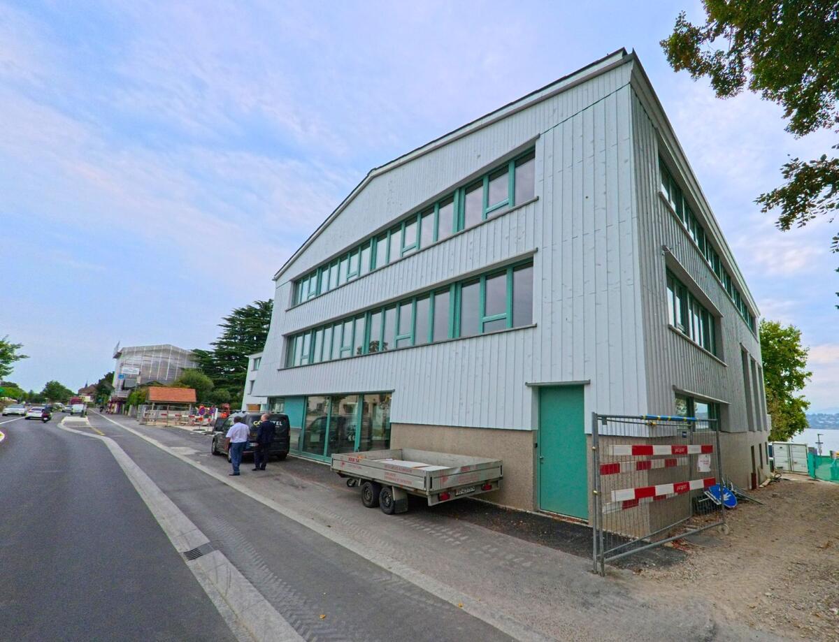 Ein modernes, helles Gebaeude mit gruenen Fensterrahmen steht an einer Strasse in Bellevue (1293, Kanton Genf), daneben sind Baustellenabsperrungen sichtbar.