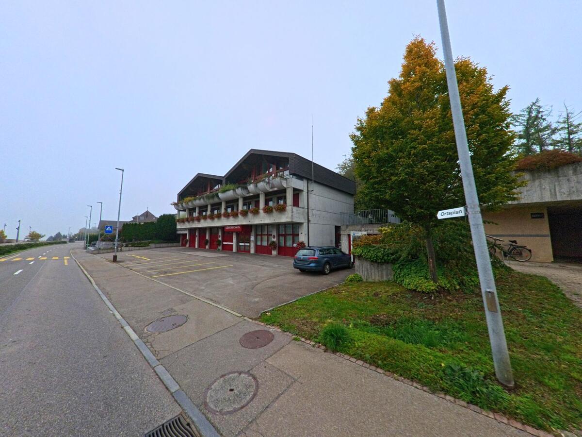 Das Feuerwehrgebäude mit roten Toren steht an einer breiten Strasse in Bellikon (5454, Kanton Aargau), umgeben von herbstlich gefärbten Bäumen.
