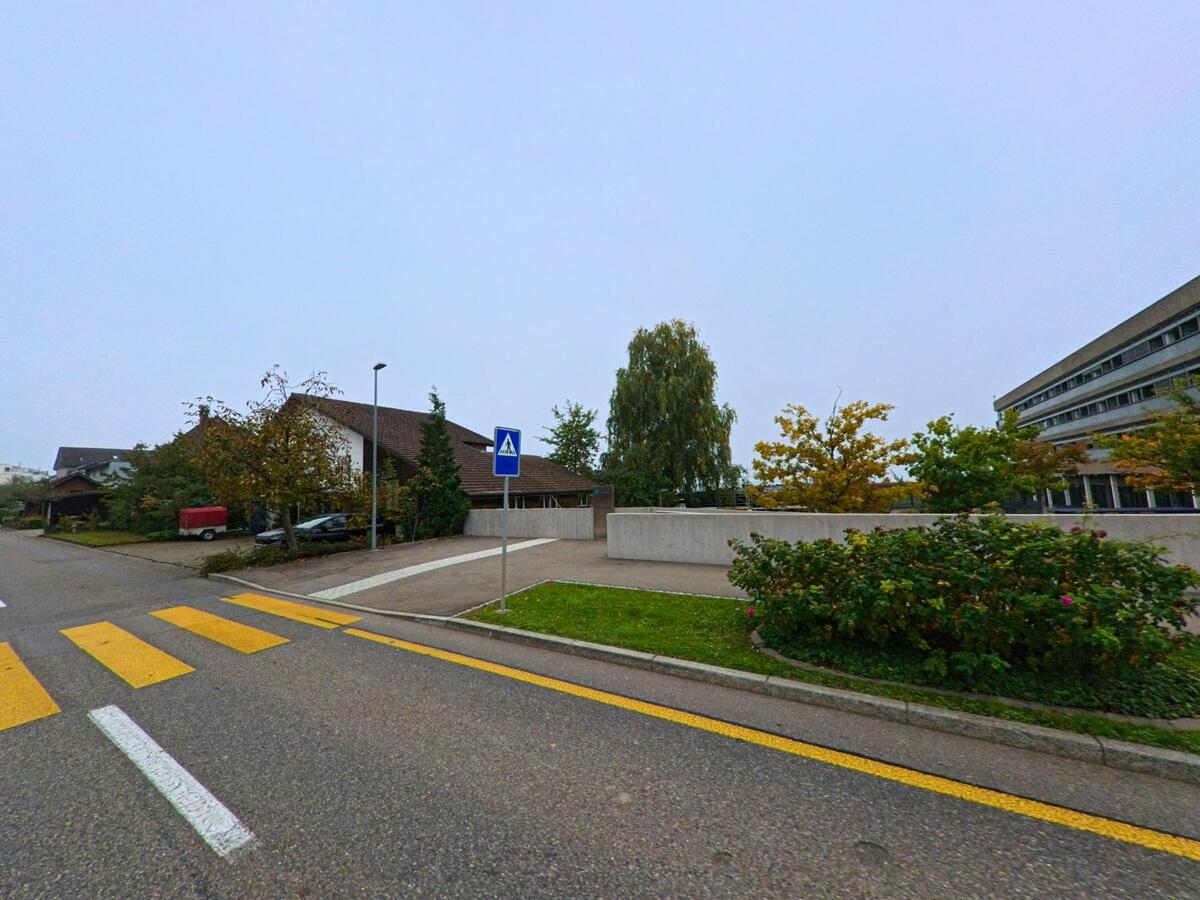Ein Fussgängerstreifen führt in Bellikon (5454, Kanton Aargau) zwischen Wohnhäusern und der Rehaklinik vorbei, umgeben von herbstlich gefärbten Bäumen.