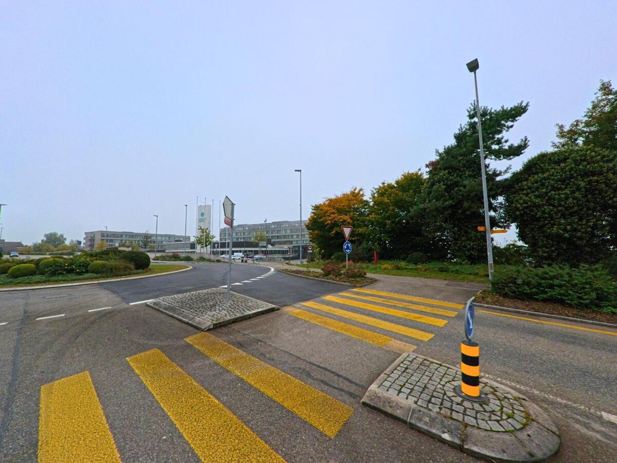 Ein Kreisverkehr mit gelbem Fussgängerstreifen und Begrünung führt zur Rehaklinik Bellikon (5454, Kanton Aargau) unter grauem Himmel.