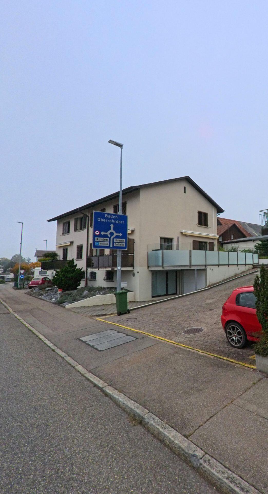 Ein modernes Wohnhaus mit Balkon steht an einer Strasse in Bellikon (5454, Kanton Aargau), neben einem Wegweiser Richtung Baden und Oberrohrdorf.