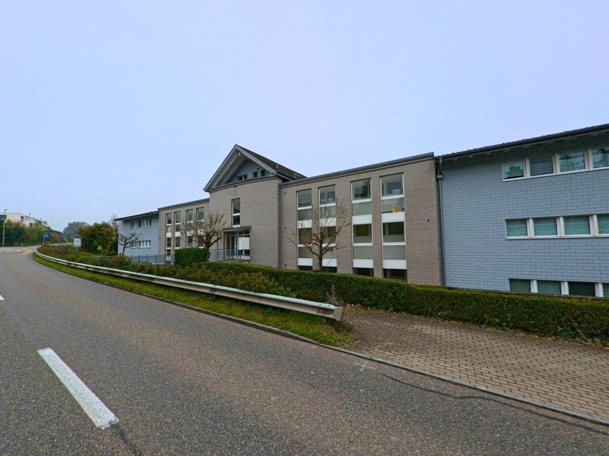 Mehrstöckige Wohngebäude mit hellen Fassaden und gepflegter Hecke säumen eine ansteigende Strasse in Bellikon (5454, Kanton Aargau).