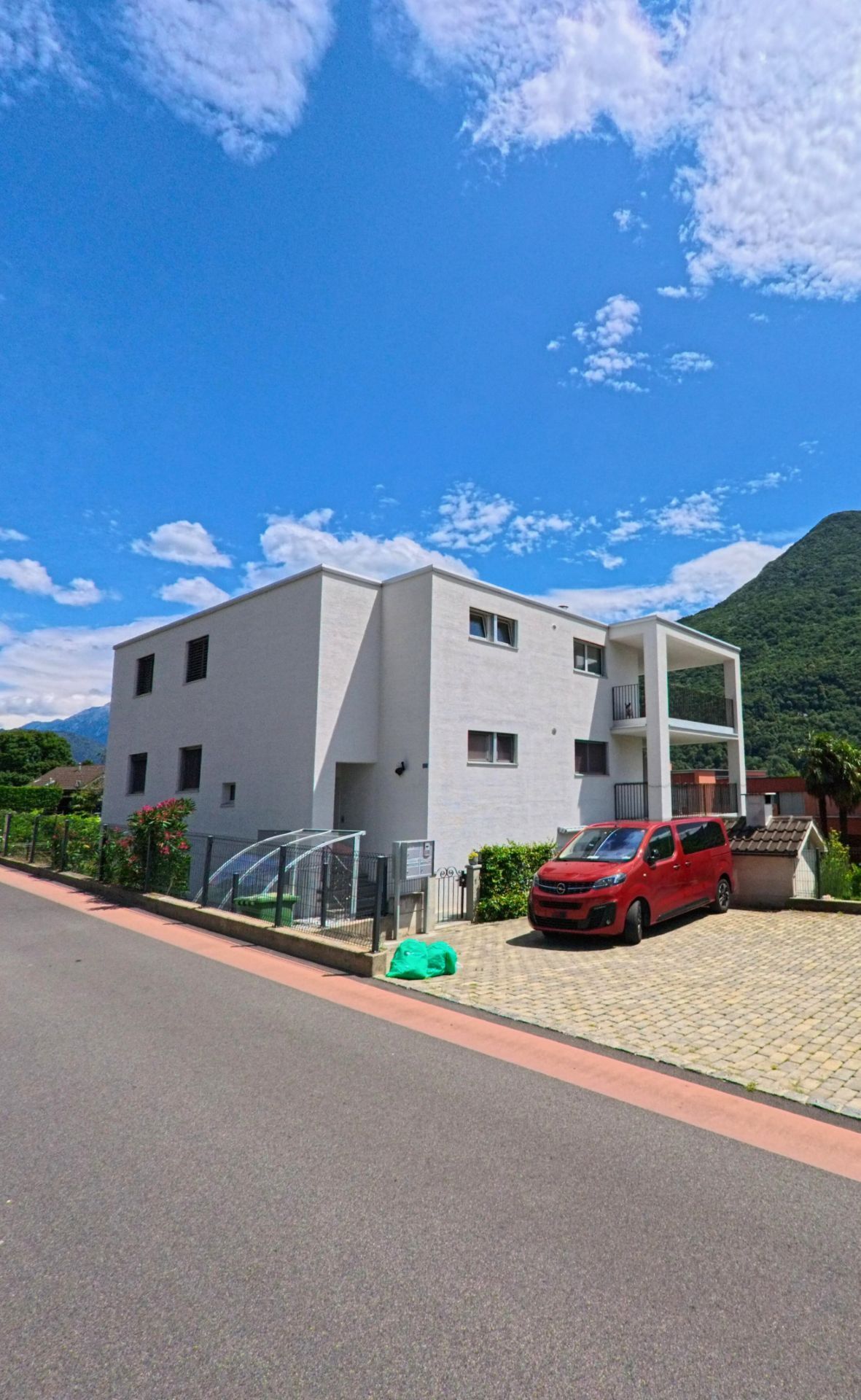 Ein modernes weisses Wohnhaus mit klaren Linien steht in Bellinzona (6503, Kanton Tessin) an einer ruhigen Strasse mit einem roten Fahrzeug auf dem Vorplatz.