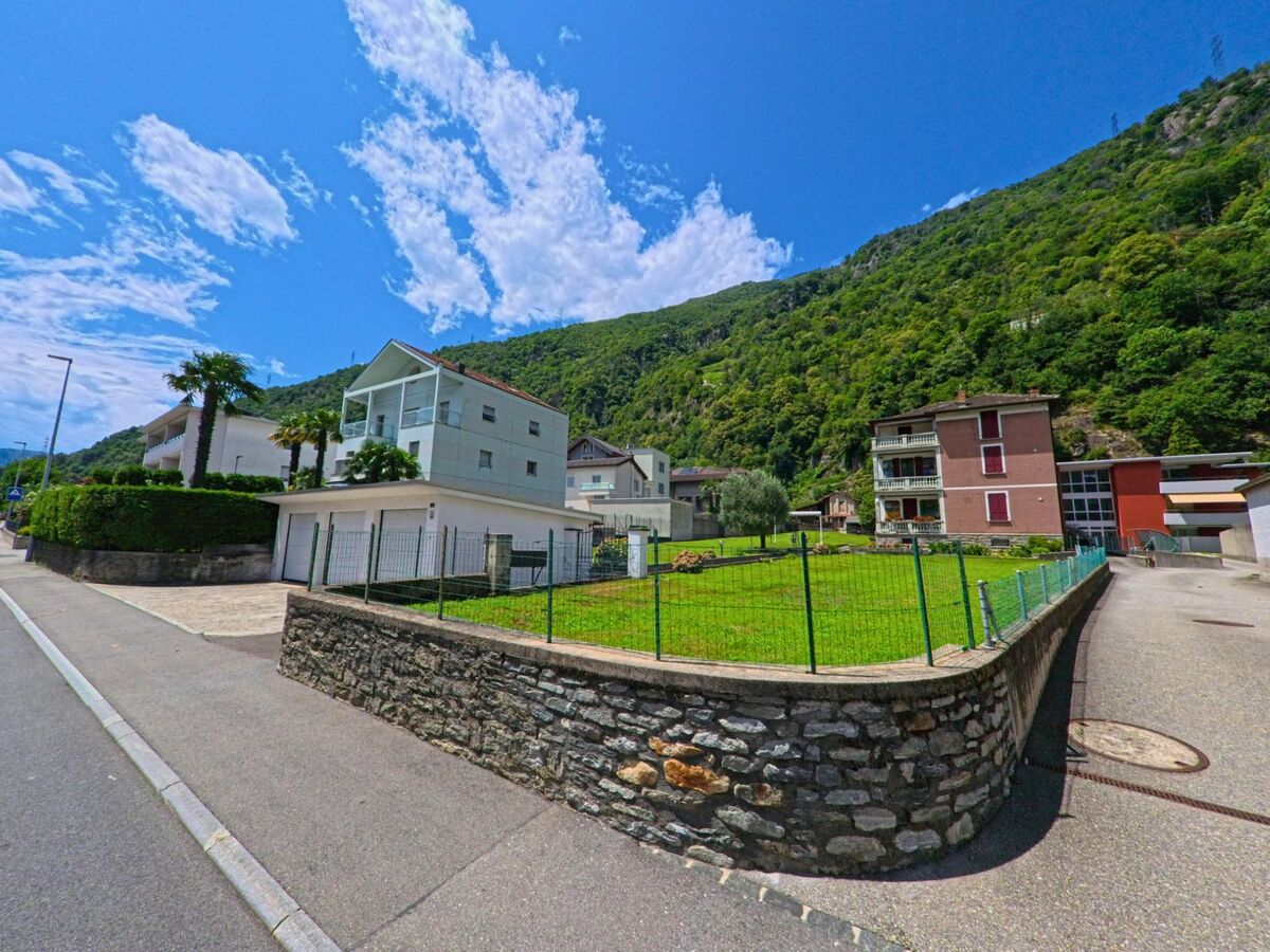 In Bellinzona (6503, Kanton Tessin) reihen sich moderne Wohnhaeuser mit Palmen und Gartenanlagen entlang einer ruhigen Strasse vor bewaldeten Berghaengen.
