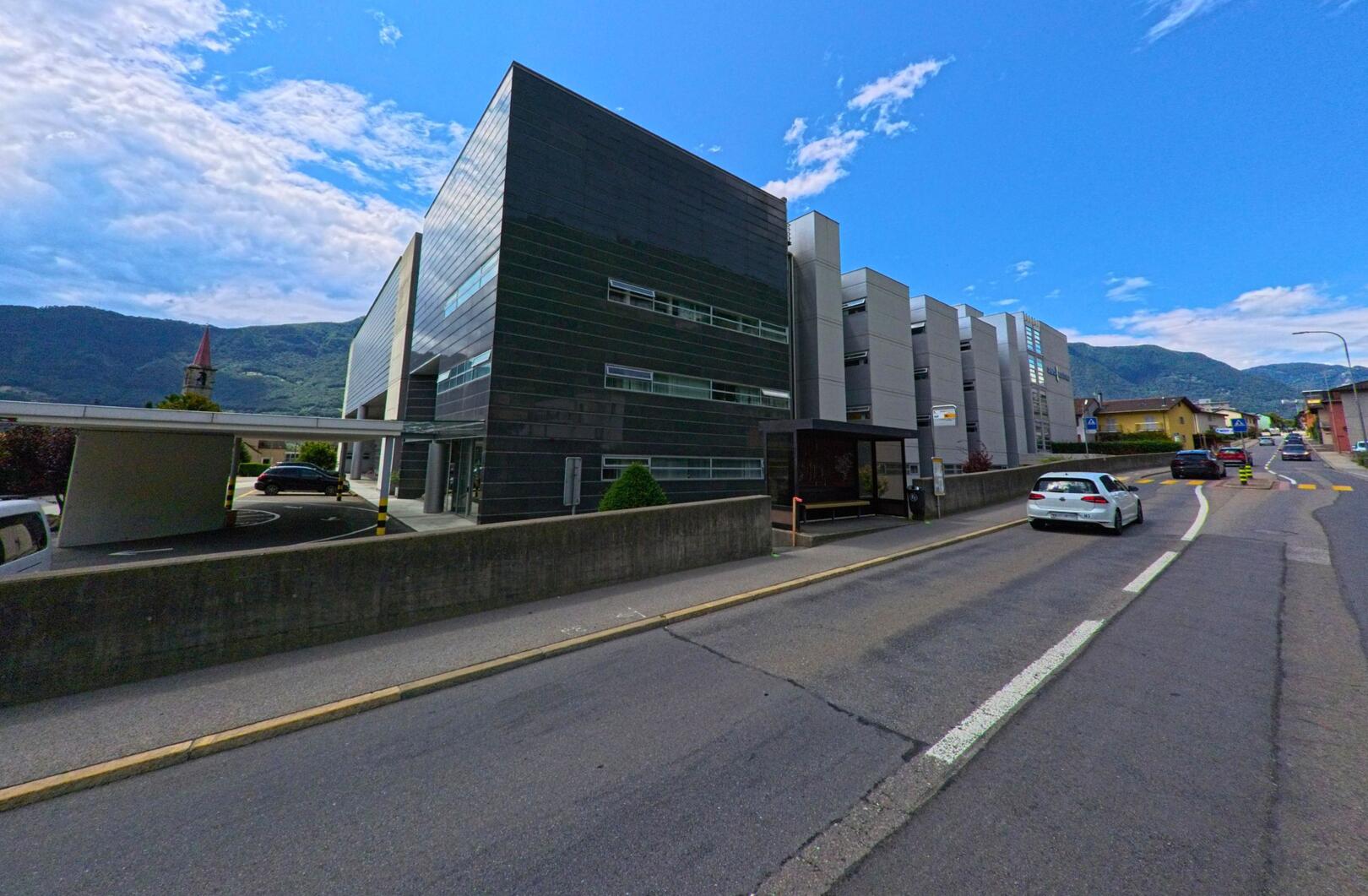 Ein modernes Gebaeude mit dunkler Fassade steht in Bellinzona (6514, Kanton Tessin) an einer befahrenen Strasse mit Blick auf die Berge im Hintergrund.