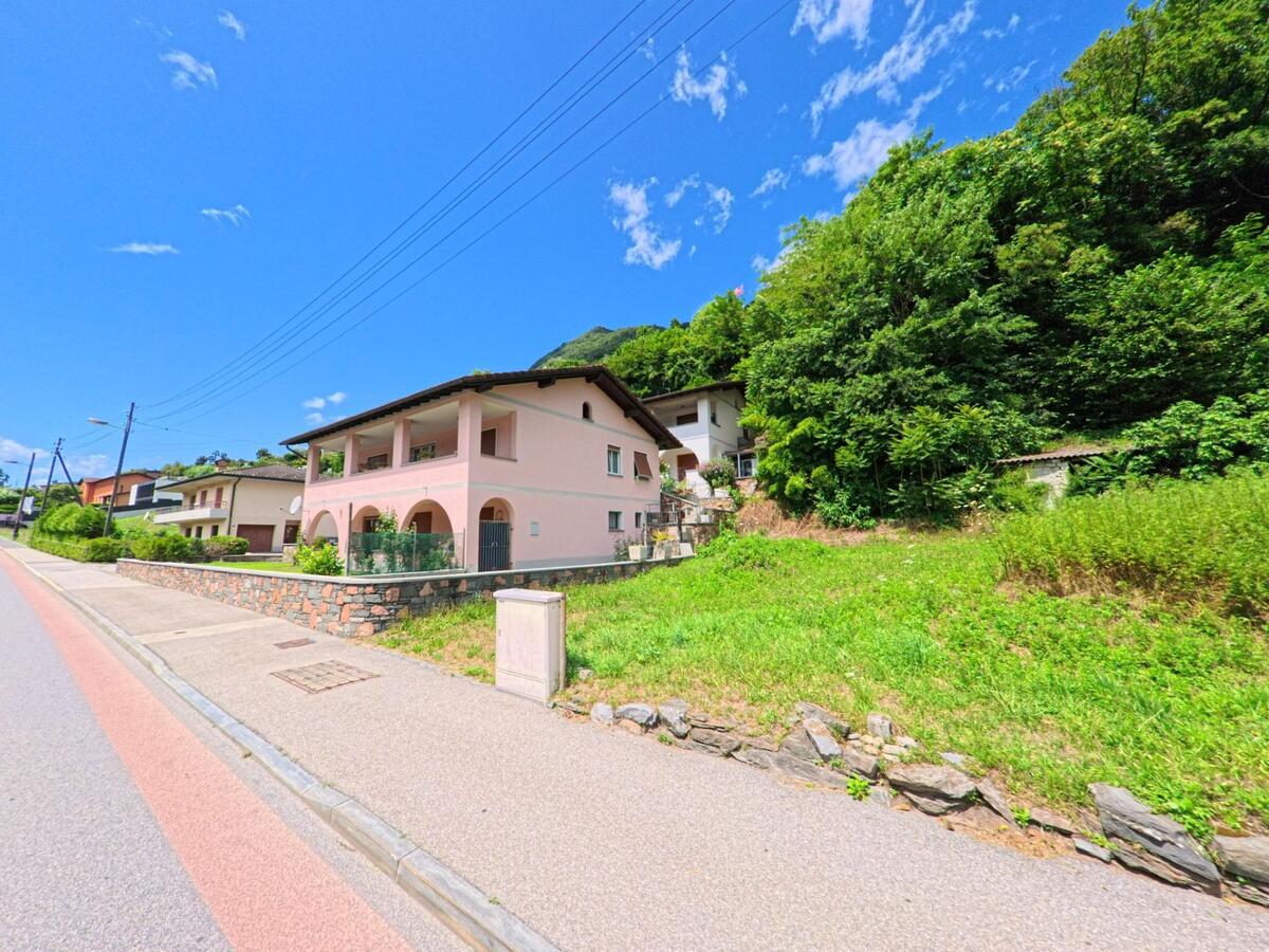 Ein rosa Wohnhaus mit grossen Rundboegen steht in Bellinzona (6515, Kanton Tessin) an einer ansteigenden Strasse, umgeben von Gruenflaechen und Baeumen.
