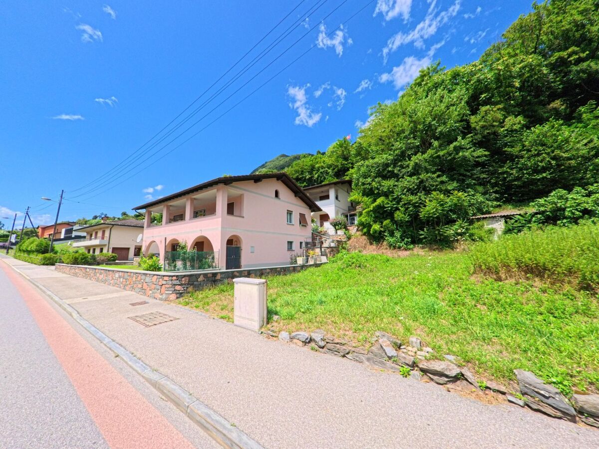 Ein rosa Wohnhaus mit grossen Rundboegen steht in Bellinzona (6515, Kanton Tessin) an einer ansteigenden Strasse, umgeben von Gruenflaechen und Baeumen.