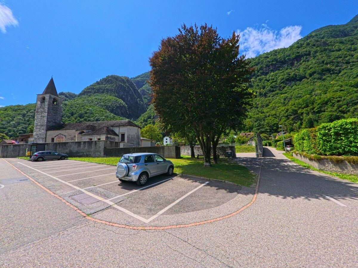 Auf einem Parkplatz in Bellinzona (6524, Kanton Tessin) stehen wenige Autos vor einer alten Kirche, umgeben von grünen Hügeln und klarem Himmel.