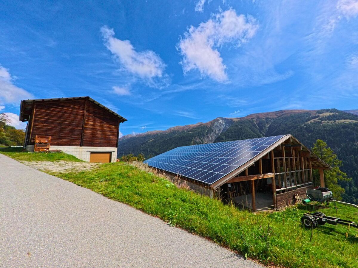Am Rand von Bellwald (3997, Kanton Wallis) steht ein Holzgebäude neben einer Scheune mit grossen Solarpanelen vor den Bergen.
