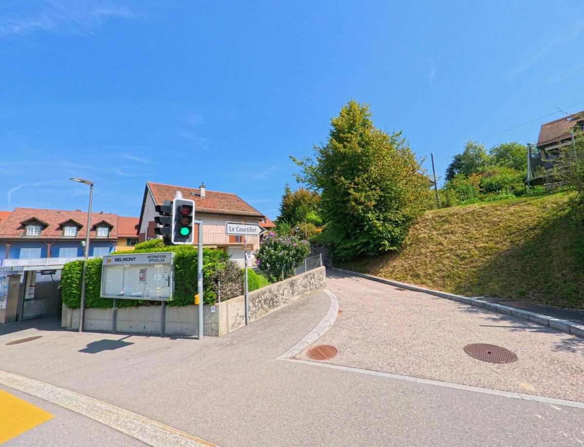 In Belmont (1092, Kanton Waadt) befindet sich eine kleine Kreuzung mit Ampel, Anschlagtafel und einem ansteigenden Weg neben Wohnhaeusern.