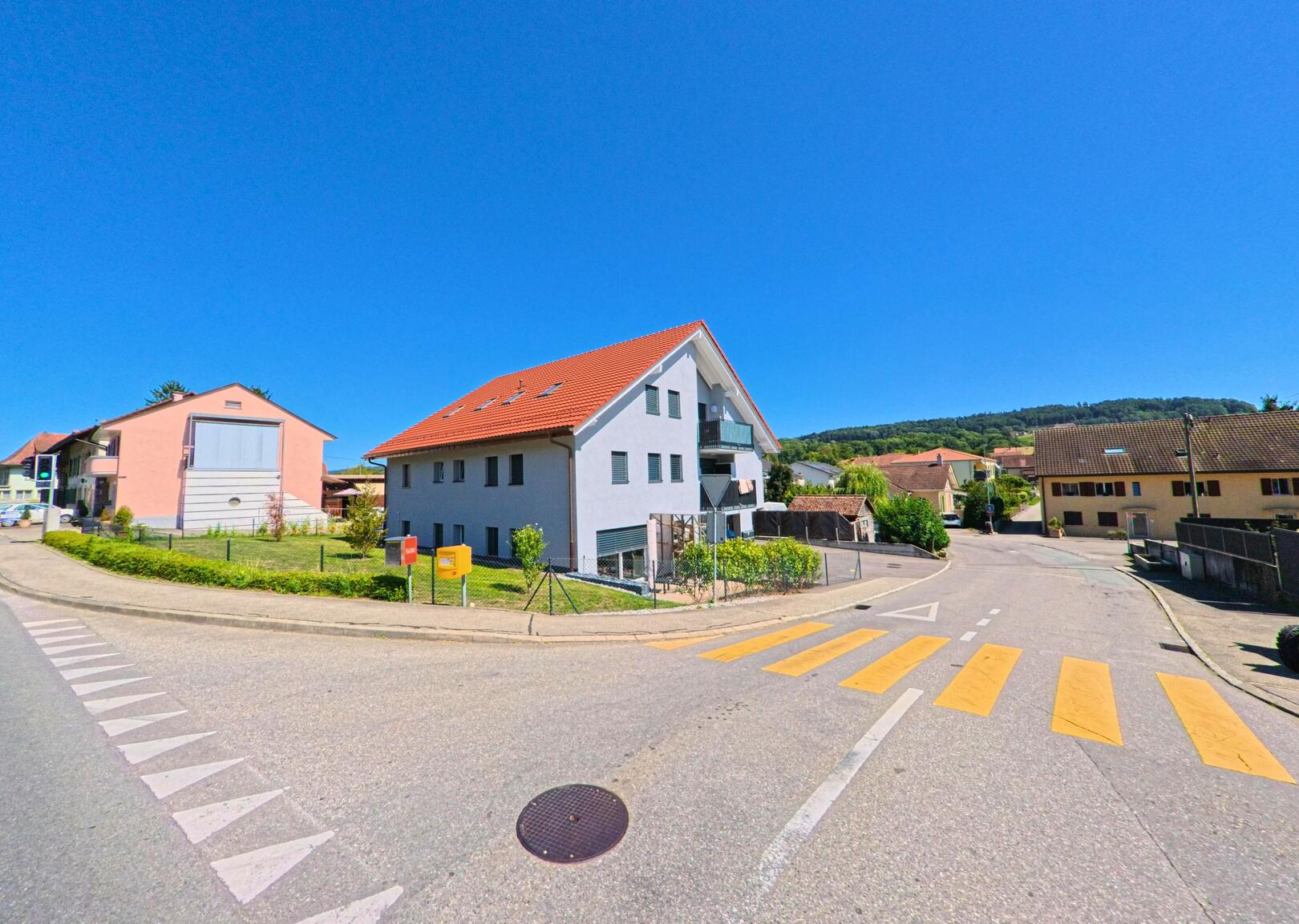 An einer Kreuzung in Belmont-Broye (1563, Kanton Freiburg) steht ein modernes Wohnhaus mit rotem Satteldach und gepflegtem Vorgarten.
