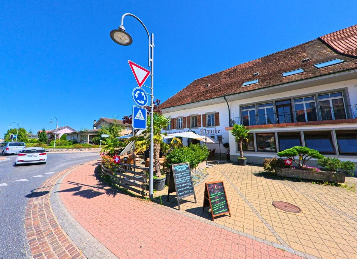 Am Rand eines Kreisverkehrs in Belmont-Broye (1564, Kanton Freiburg) befindet sich ein Restaurant mit Aussenterrasse, umgeben von Palmen und dekorativen Pflanzen.