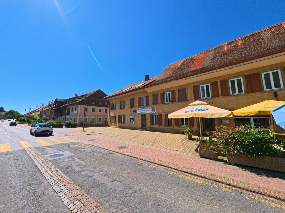 An einer sonnigen Hauptstrasse in Belmont-Broye (1564, Kanton Freiburg) befinden sich ein Restaurant mit Aussenterrasse und ein Immobilienbuero in traditionellen Gebaeuden.