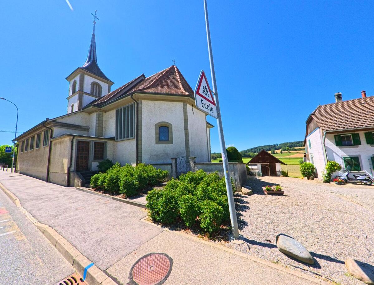 In Belmont-Broye (1773, Kanton Freiburg) steht eine Kirche mit hohem Turm und Spitzdach an einer Strasse, neben der sich ein Hof mit Wohnhaus und Blick auf Felder erstreckt.