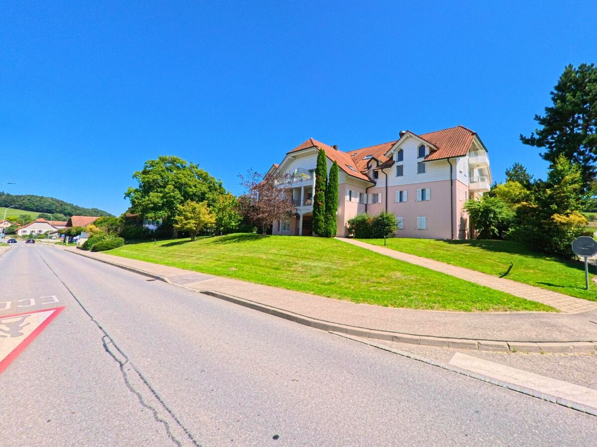 An einer breiten Strasse in Belmont-Broye (1773, Kanton Freiburg) steht ein grosses Wohngebaeude mit hellrosa Fassade und gepflegtem Gruenbereich.