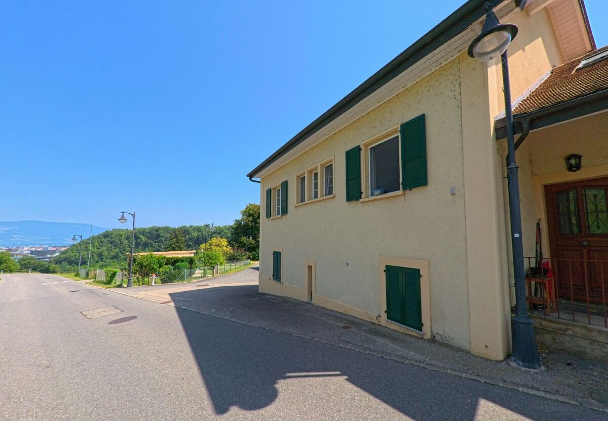 Ein Haus mit gruenen Fensterlaeden steht in Belmont-sur-Yverdon (1432, Kanton Waadt) direkt an einer abfallenden Strasse mit Blick ins Tal.