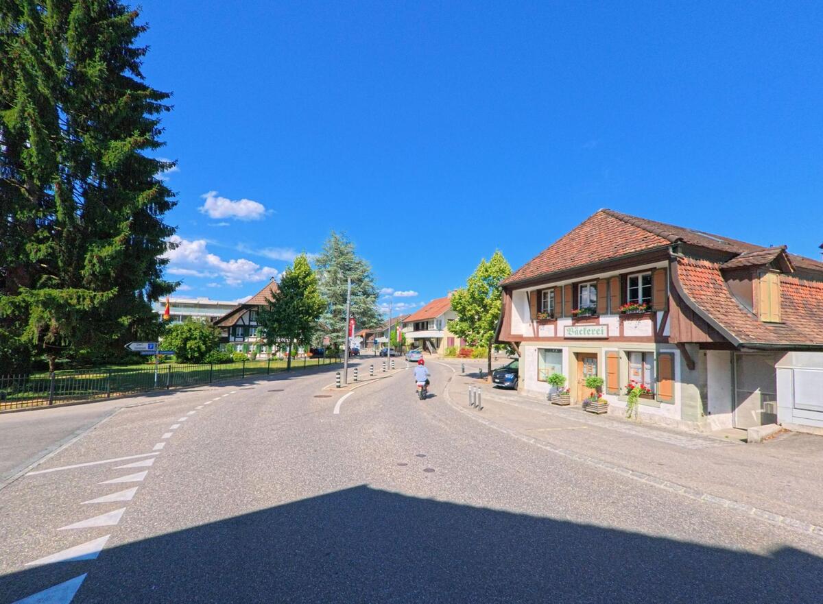 In Belp (3123, Kanton Bern) liegt eine Bäckerei mit rotbraunem Ziegeldach an einer leicht geschwungenen Strasse, gegenüber einem grossen Nadelbaum und nahe dem Dorfzentrum.